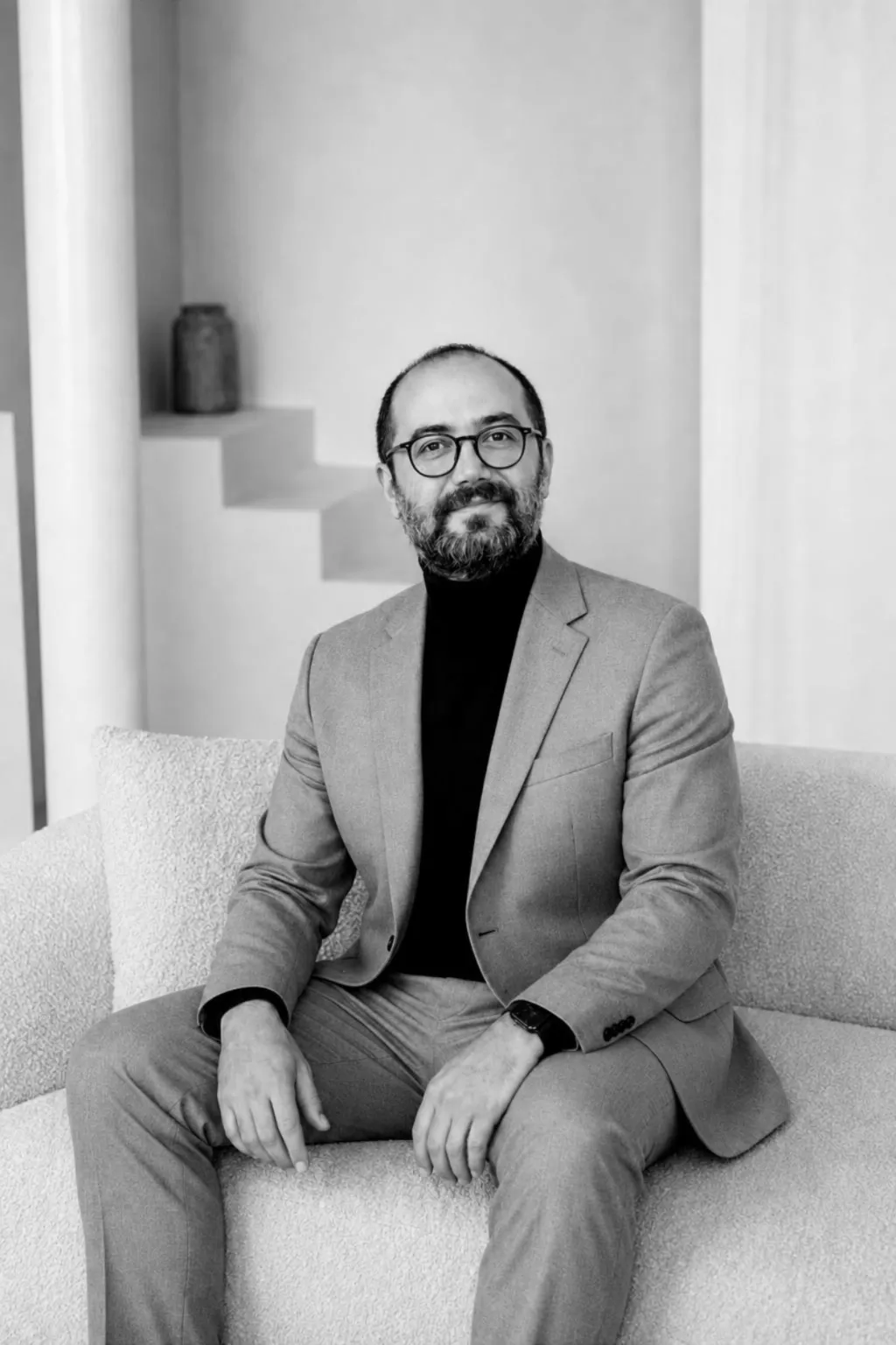 A man with glasses and a short beard sits on a boucle sofa in a light grey suit over a black turtleneck, hands resting on his knees — black and white.