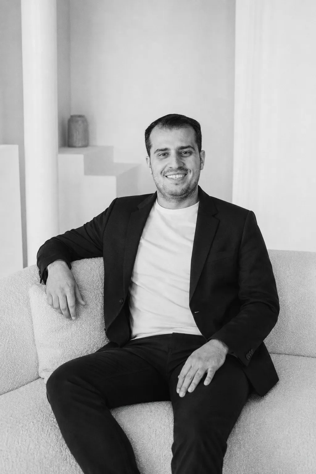 A man with short dark hair sits on a boucle sofa in a dark blazer over a white t-shirt, smiling, one arm resting along the back of the sofa — black and white.
