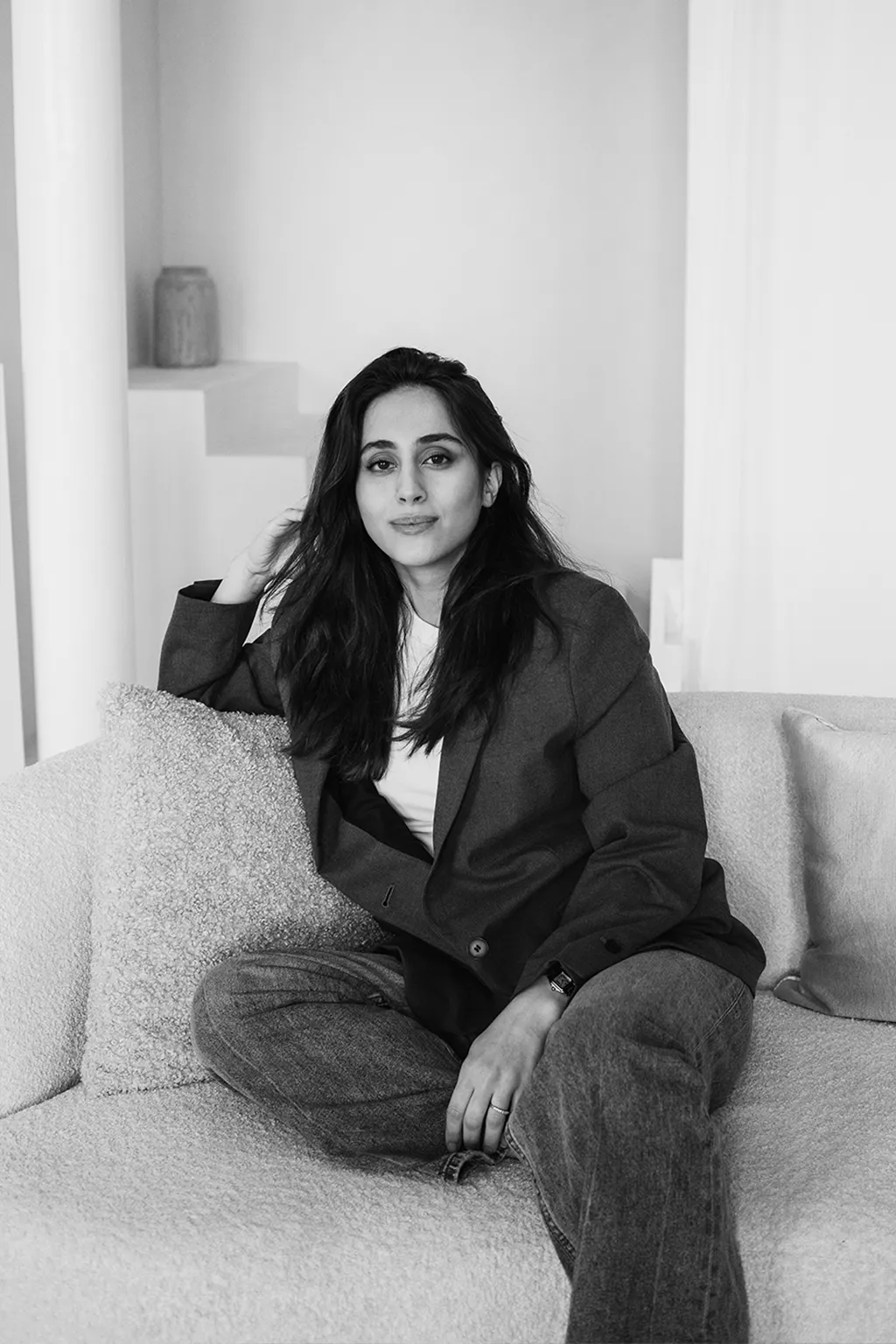 A woman with long dark hair sits cross-legged on a boucle sofa in an oversized blazer, one hand resting near her hair — black and white.